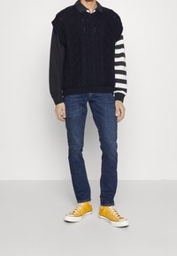 Pull sans manches tricoté bleu marine sur une chemise à manches longues noire avec des rayures blanches, associé à un jean bleu foncé et des baskets jaunes.