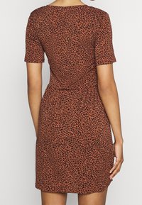 Oranssi-ruskea lyhythihainen mekko mustalla leopardikuosilla, vartaloa myötäilevä siluetti ja taakse kiristettävä yksityiskohta. Pehmeä, teksturoitu kangas.