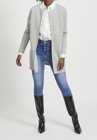 Kvinna klädd i en ljusgrå strukturerad öppen jacka, vit skjorta med knappar, blå tajta jeans och svarta knähöga stövlar.