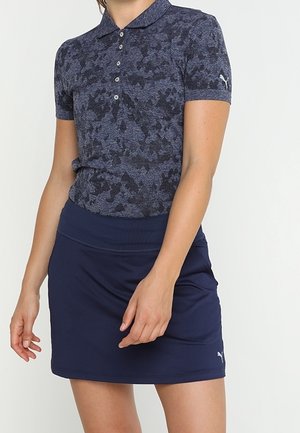 Kvinde iført en mørkeblå mønstret polo-shirt og en ensfarvet marineblå nederdel, begge med Puma-logoer, stående mod en ensfarvet baggrund.