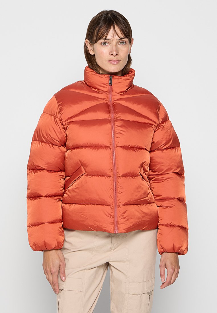 Eine Frau trägt eine glänzende orange Puffjacke mit hohem Kragen und beigen Hosen und steht vor einem einfarbigen, hellen Hintergrund.