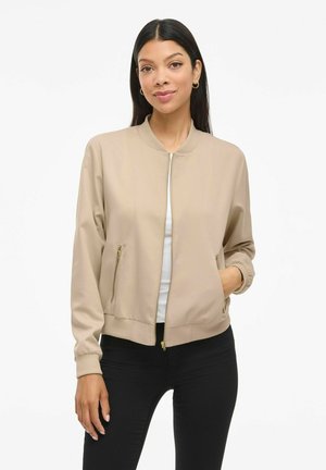 Lichtbeige bomberjack met ritssluiting aan de voorkant, ribbels aan de mouwen, twee met rits afgesloten zakken en een gladde textuur. Draagt over een witte top en zwarte broek.