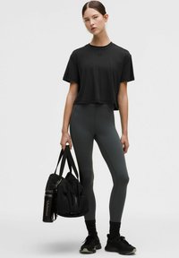 Fekete, rövid fazonú póló és magas derekú, szürke leggings. A modell egy fekete táskát tart, benne egy vizespalackkal; a leggings oldalán csíkok láthatók, sima textúrával.