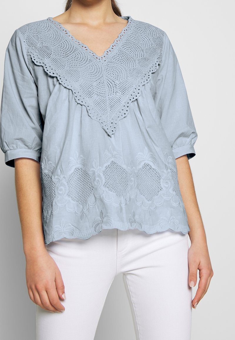 Světle modrý top s výstřihem do "V" z háčkované krajky a květinovou výšivkou podél lemu. Jemná tkanina s nabíranými rukávy.