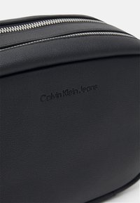 Černá toaletní taška z imitace kůže s texturovaným povrchem, která má stříbrný zip a embosované logo "Calvin Klein Jeans".