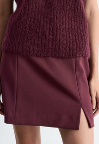 Bordeauxfarbener Wollmischung-Westen ohne Ärmel mit strukturiertem Rippenstrick, kombiniert mit einem taillierten bordeauxfarbigen Minirock mit seitlichem Schlitz.