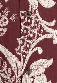 Tessuto bordeaux con motivo floreale bianco, dotato di chiusura a bottone. La trama testurizzata con una leggera irregolarità aggiunge profondità al design.