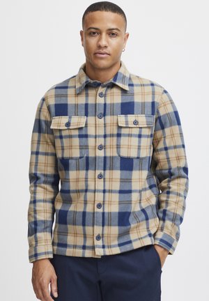 Chemise en flanelle à carreaux beige et bleu avec deux poches poitrine, fermeture à boutons et manches longues. Texture douce avec des motifs contrastés.