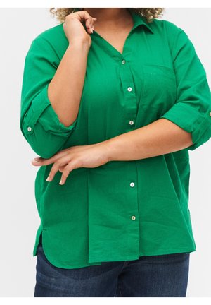 Grünes Hemd zum Knöpfen mit einer Brusttasche, hochgekrempelten Ärmeln und einem lockeren Schnitt. Glatte Textur mit kleinen, runden Knöpfen und Seitenschlitzen.
