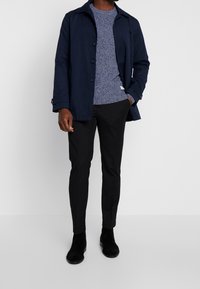 Manteau bleu marine avec un col, fermeture à boutons, superposé sur un pull bleu texturé, associé à un pantalon noir ajusté et des bottes en daim noir.