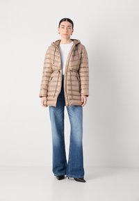 Giacca imbottita beige con cappuccio, vita con cordoncino e due tasche laterali con zip, indossata sopra una maglietta bianca e abbinata a jeans blu a zampa d'elefante.