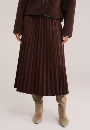 Vrouw draagt een bruine geplooide midi rok, een gestructureerd bruin jasje en beige suède kniehoge laarzen, staand op een neutrale achtergrond.