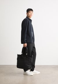 Zwarte stoffen tote bag met dubbele handvatten en zilveren hardware, gedragen met een donker shirt en donkere broek. Zachte textuur, functioneel ontwerp.