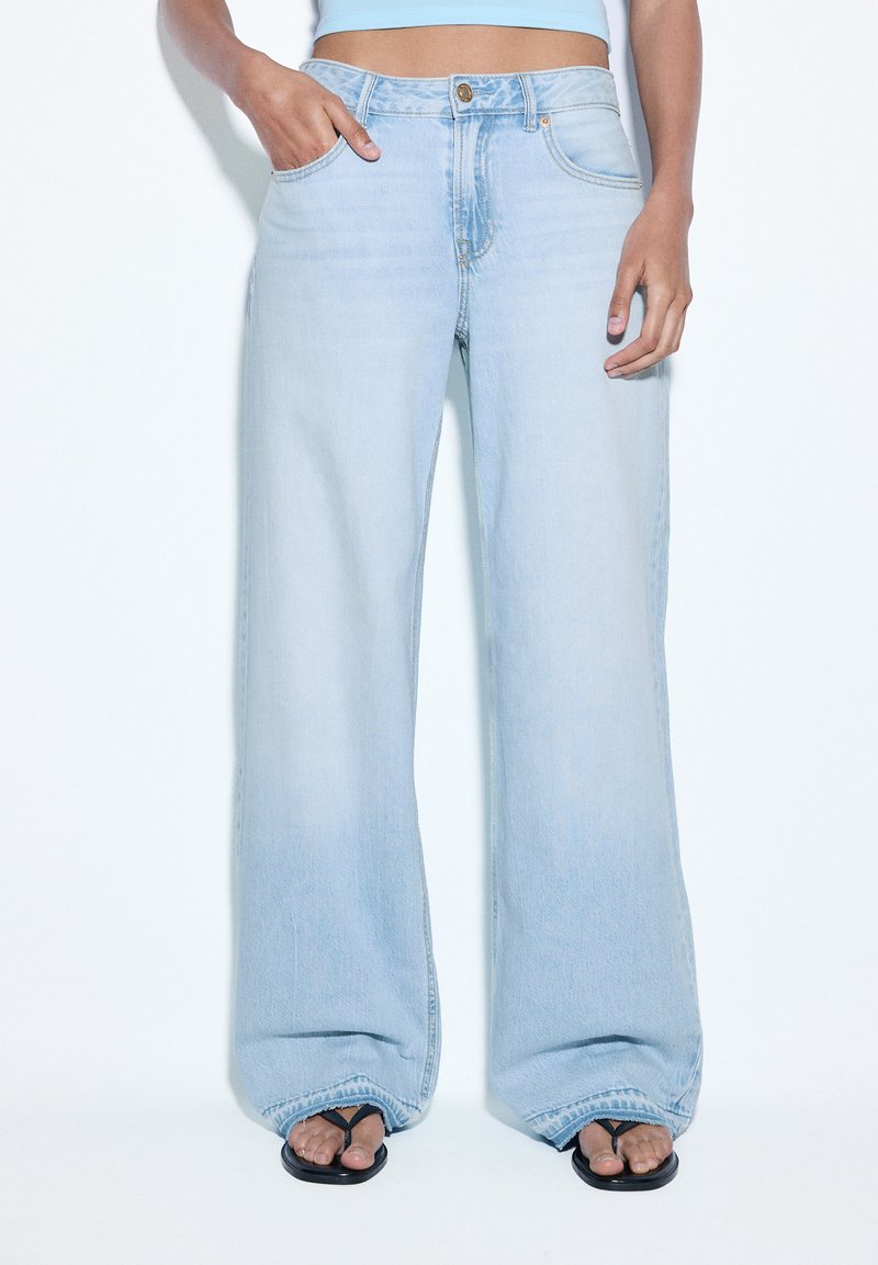 Jean bleu clair à jambes larges porté avec des tongs noires, une main dans la poche, top bleu clair court laissant voir le ventre.