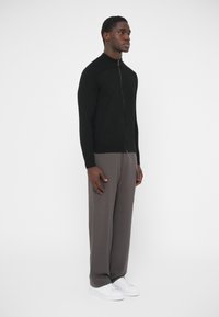 Čierny pletený kardigán na zips s rebrovanými akcentmi na ramenách, kombinovaný so sivými širokými nohavicami a bielymi teniskami, na neutrálnej pozadí.