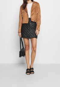 Donna che indossa una giacca in suede marrone, un top bianco, una gonna in pelle trapuntata nera, sandali neri e tiene una borsa trapuntata nera.