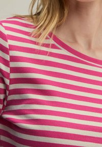 Vrouw in een geribbelde trui met horizontale roze en witte strepen, die haar nek en blonde haar toont op een lichte achtergrond.