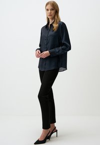 Marineblauwe blouse met knopen en een ruitpatroon, gecombineerd met zwarte slim-fit broek en zwarte puntige hakken. Gladde stoftextuur.