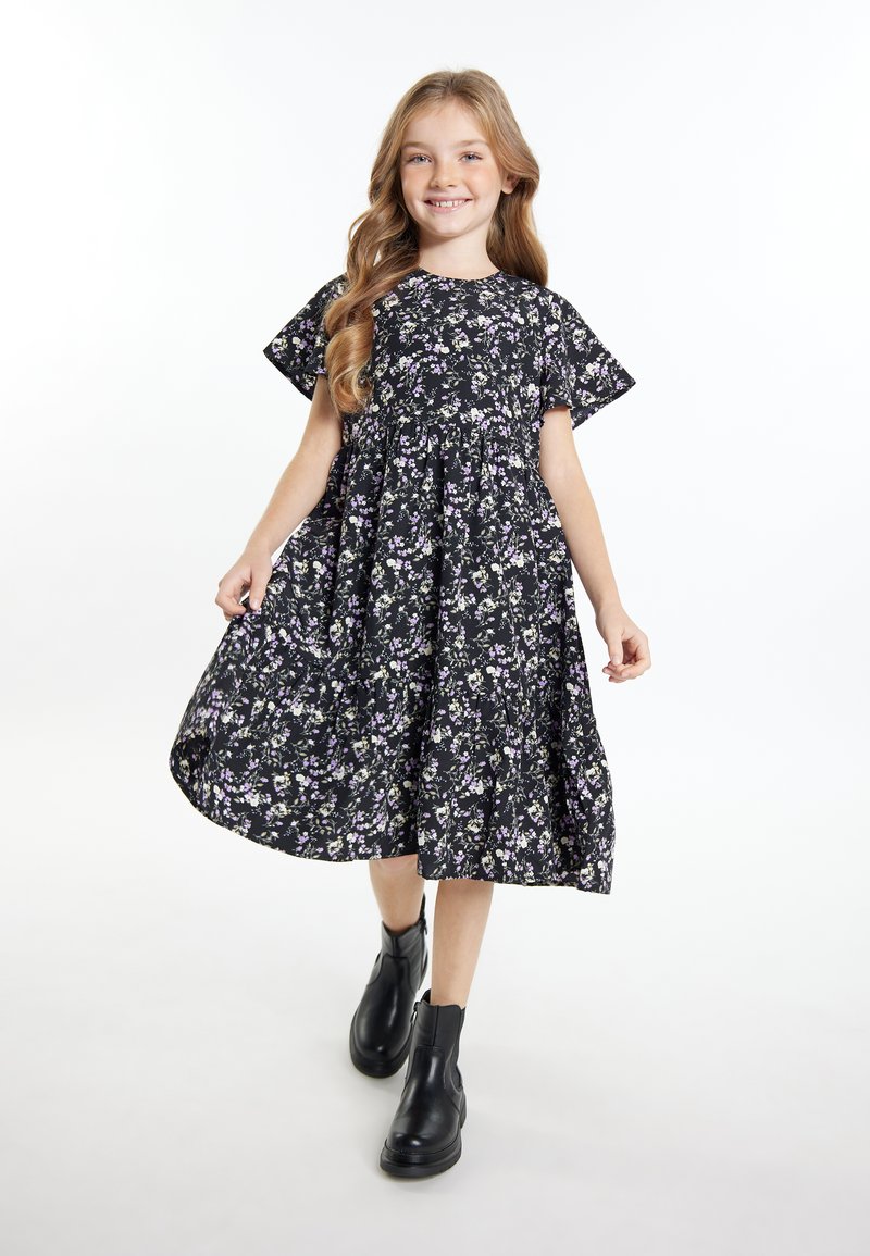 Zwart bloemenjurk met korte mouwen, voorzien van paarse en witte bloemen, geplooide taille en vloeiende rok. Gekozen in combinatie met zwarte enkellaarsjes.