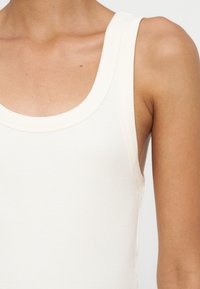 Gros plan d'une personne portant un débardeur blanc côtelé sans manches, montrant l'épaule, le haut du bras et la zone de la clavicule.