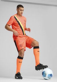 Equipación de fútbol naranja con una franja horizontal negra y amarilla, combinada con botas negras y calcetas naranjas, junto a un balón de fútbol azul y blanco.