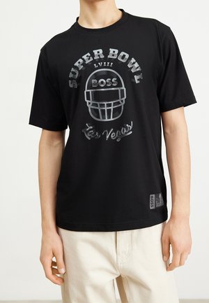 Henkilö, joka on pukeutunut mustaan t-paitaan, jossa on teksti "SUPER BOWL LVIII", jalkapallokypärän grafiikka ja teksti "Las Vegas", yhdessä vaaleanväristen housujen kanssa.