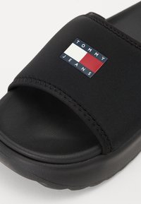 Černé skluzové sandály s texturovanou gumovou podrážkou. Mají široký pásek vyrobený z hladké tkaniny a obdélníkové logo v červené, bílé a modré barvě.