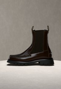 Bottine en cuir marron foncé avec panneaux élastiques noirs sur les côtés, languettes, couture moc toe et une épaisse semelle crantée noire sur une surface neutre.
