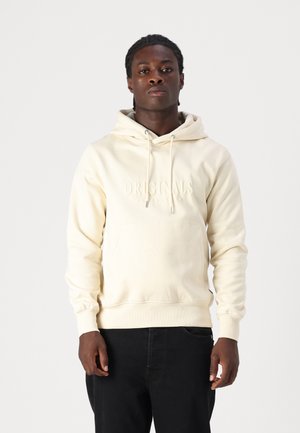 Crème hoodie met "ORIGINALS" in reliëf op de voorkant, voorzien van een capuchon met trekkoord en een buidelzak, gecombineerd met zwarte broek.