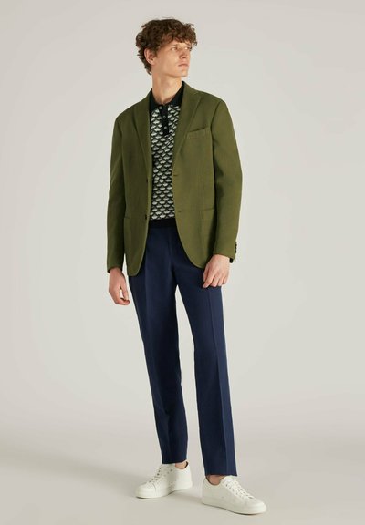 Blazer vert olive sur un polo noir et blanc à motifs, accompagné de pantalons navy et de baskets blanches. Coupe ajustée, lignes épurées.