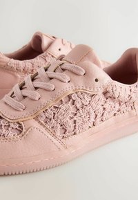 Des baskets roses présentent des détails en tissu texturé, un cuir synthétique lisse, une forme de bout arrondi, un design à lacets à plat et une semelle en caoutchouc avec des bords surélevés.
