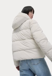 Een lichtbeige pufferjas met horizontale gestikte gedeeltes, een capuchon en elastische manchet, gecombineerd met lichtblauwe high-waisted jeans.