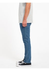 Jeans de mezclilla ajustados de color azul combinados con una camiseta gris, llevados con zapatillas de lona en blanco y negro, mostrados desde el lado.