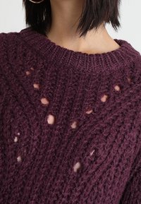 Maglione lavorato a maglia in un profondo viola con un motivo testurizzato che presenta dettagli a traforo e un colletto a coste. Costruzione accogliente in filato spesso.