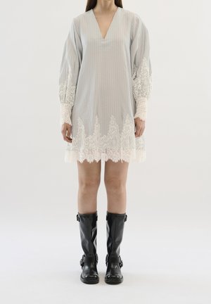 Mujer con vestido de manga larga gris claro con ribete de encaje blanco y botas de cuero negras hasta la mitad de la pantorrilla, de pie frente a un fondo sencillo.