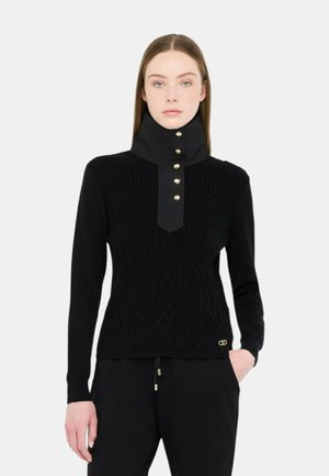 Maglione nero a maglia con colletto alto, dettagli in bottone dorato e motivi a diamante a rilievo. Indossato con pantaloni jogger neri.
