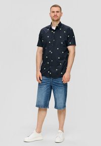 Korte mouwen marineblauw shirt met een groen sterrenpatroon, gecombineerd met blauwe denim korte broek. Model draagt witte sneakers.