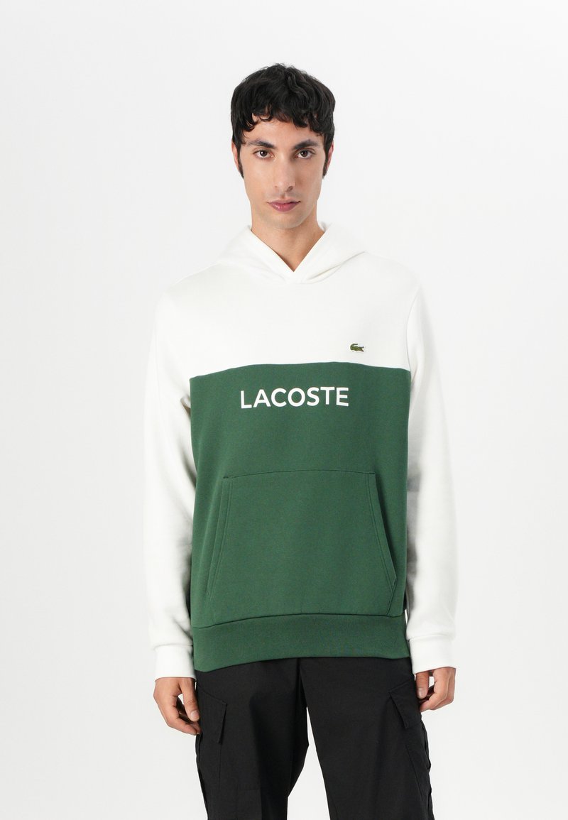 Roheline ja valge kapuutsiga sviiter, millel on känguru-tasku, millel on rinnal valge print "LACOSTE" ja väike logo vasakul.