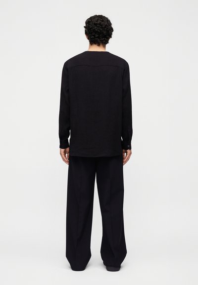 Άτομο με σγουρά σκουρόχρωμα μαλλιά στέκεται γυρισμένο προς τα πίσω, φορώντας χαλαρό μαύρο μακρυμάνικο πουκάμισο και φαρδιά μαύρα παντελόνια μπροστά σε απλό φόντο.