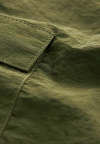 Material de culoare verde măsliniu cu o suprafață texturată, având cusături vizibile și un detaliu cu pliu. Apare neted și ușor șifonat.