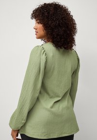 Blouse verte texturée à manches longues avec des accents de épaules froncés, présentant une coupe décontractée et des détails subtils sur le tissu.