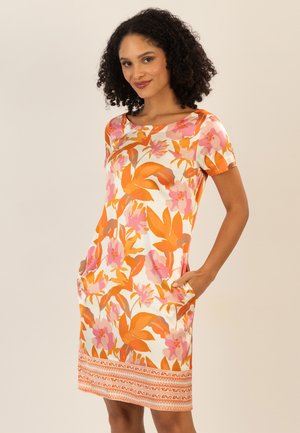 Frau trägt ein kurzärmeliges, knielanges Kleid mit orange-rosa Blumenmuster, Hände in den Taschen, steht vor einem einfarbigen Hintergrund.