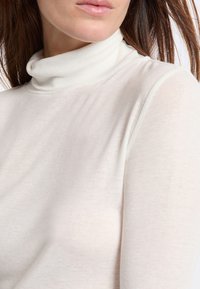 Primo piano di una donna che indossa un maglione bianco a maniche lunghe e dolcevita, mostrando le sue labbra, collo e parte superiore del torso.