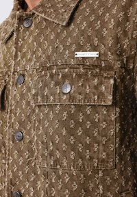 Veste marron texturée avec des boutons en métal et une poche à rabat, présentant une petite étiquette en métal "PROJECT X PARIS" au-dessus de la poche.