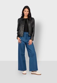 Černá kožená bundu na zip s límečkem, spárovaná s vysokými modrými džíny s širokými nohavicemi a světlými teniskami.