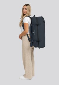 Sac à dos bleu foncé avec des bretelles réglables, une fermeture éclair et des accents de logo, porté sur le dos d'une personne. Matériau lisse et durable.