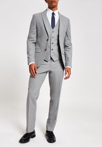 Grauer Dreiteiler mit Reverskragen, taillierte Passform, Weste mit fünf Knöpfen, kombiniert mit einem weißen Hemd und einer blauen Krawatte, ergänzt durch glänzende schwarze Schuhe.