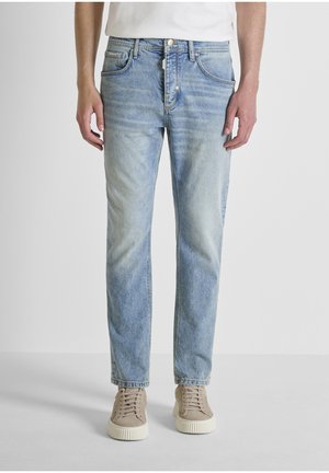 Vaqueros rectos de color azul claro desgastado, combinados con zapatillas beige y una camiseta blanca, de pie contra un fondo gris liso.