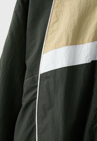 Gros plan sur un tissu montrant une veste avec des blocs de couleur vert foncé, blanc et beige et des détails de couture visibles.