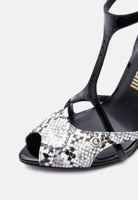 Μαύρο σανδάλι με τακούνι από δερμάτινο λουστρίνι, με λουράκι στα δάχτυλα με μοτίβο λευκού και μαύρου δέρματος φιδιού και μικρή χρυσή κυκλική λεπτομέρεια στο πλάι.